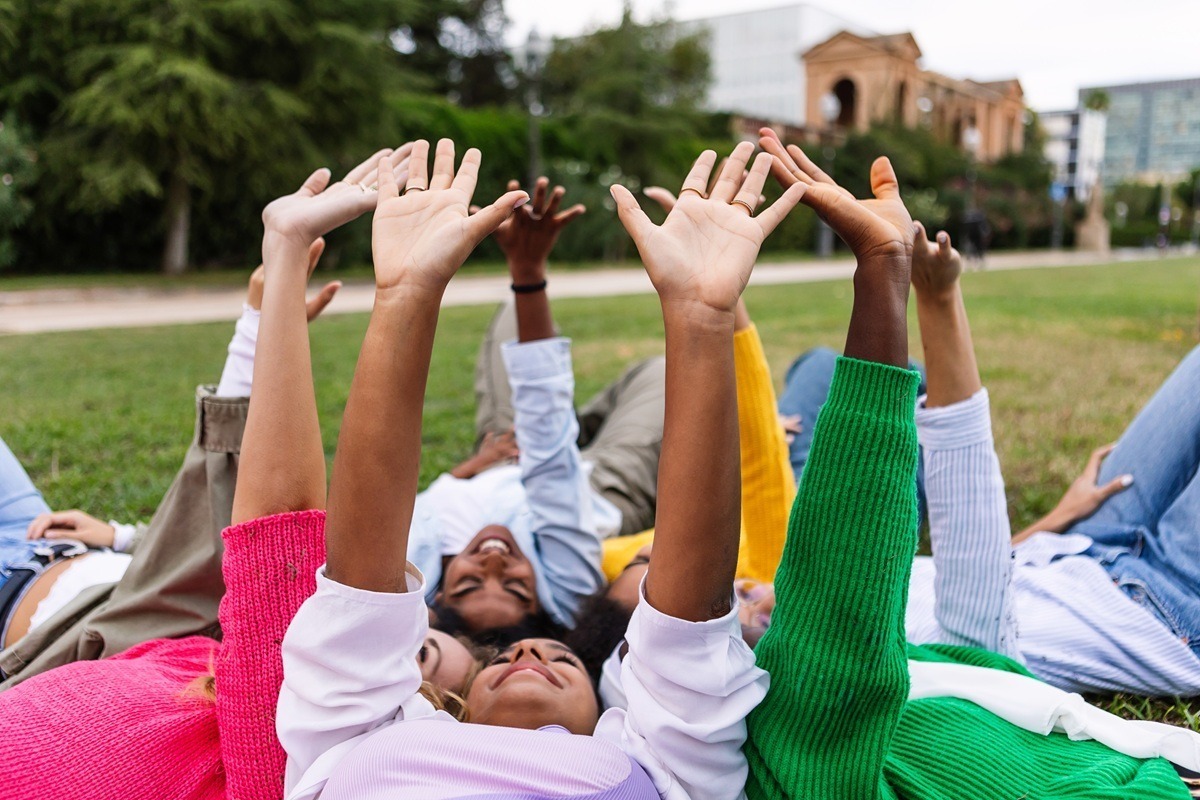 United group of diverse millennial women raising hands to the sky while lying together in circle at city park. Hope, support and volunteer community concept.