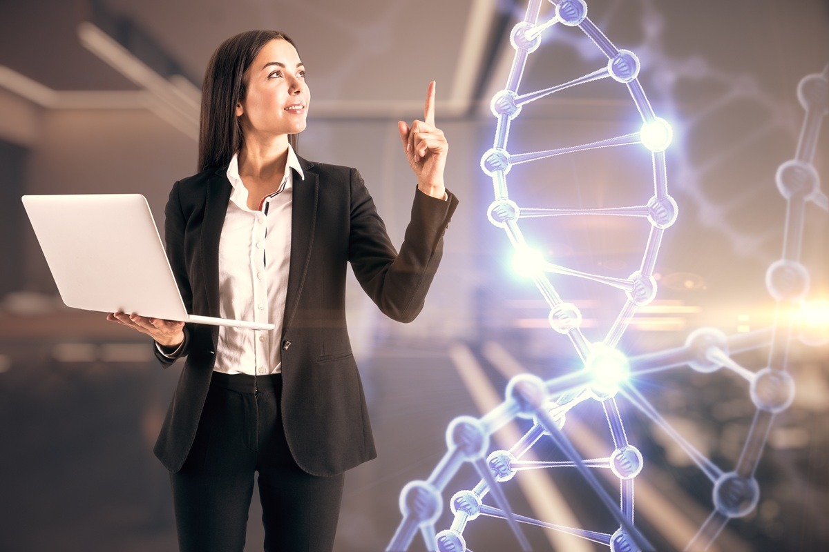 A woman wearing a black blazer and holding a white laptop poses in front of a luminous depiction of a DNA helix.