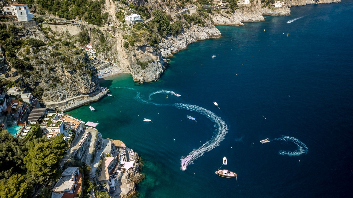 An aerial view of the blue sea against rocky Amalfi coast covered with greenery and buildings. An aerial view of the blue sea against rocky Amalfi coast covered with greenery and buildings.
