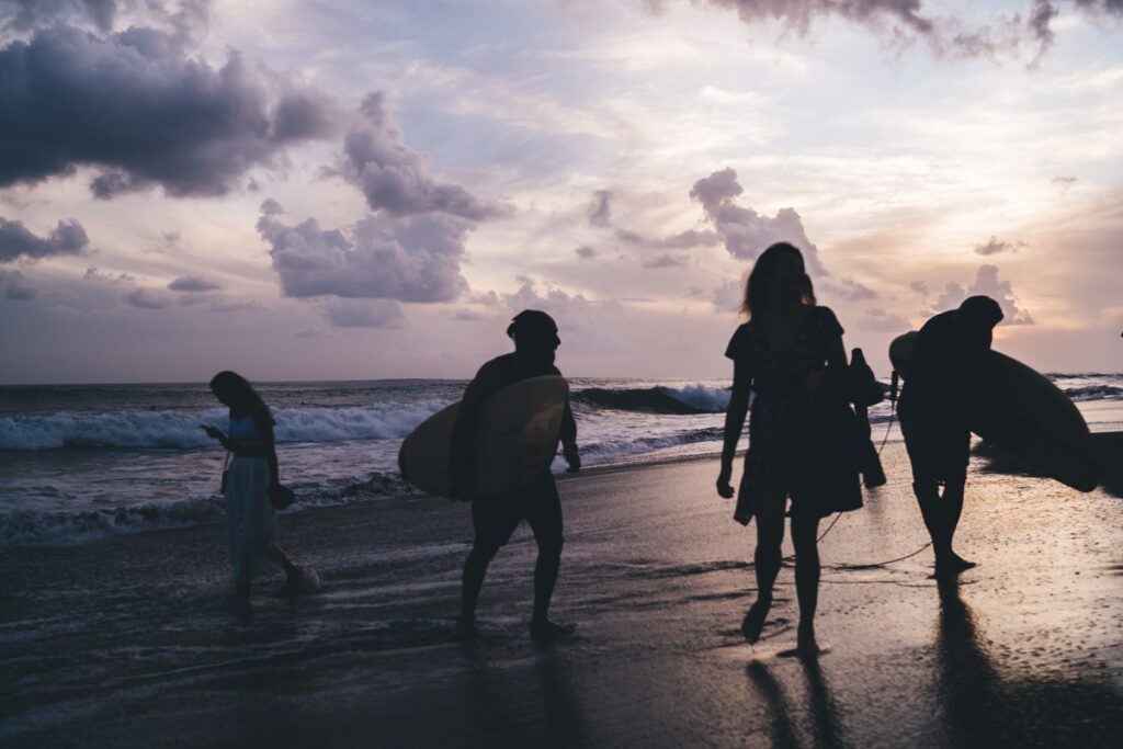 Silhouette view of young people in wetsuits walking along evening seashore holding surfboards, unrecognizable male and female surfers communicate near ocean holding boards - summertime hobby vacations