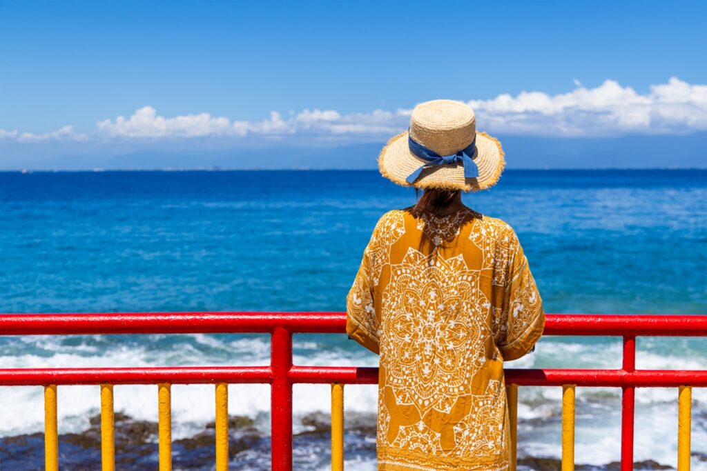 Woman in a straw hat and patterned yellow cover-up stands by a red railing, gazing at the blue ocean under a clear sky with distant clouds.