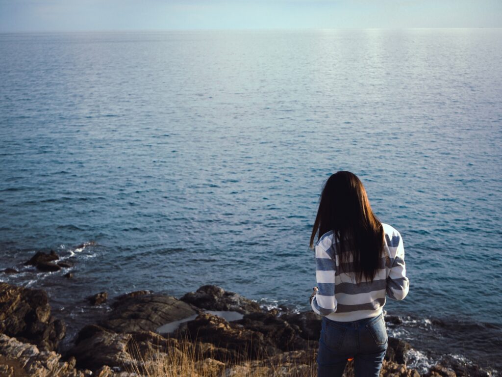 A woman overlooks the ocean from a cliff, surrounded by rocky terrain and a bright sky.
