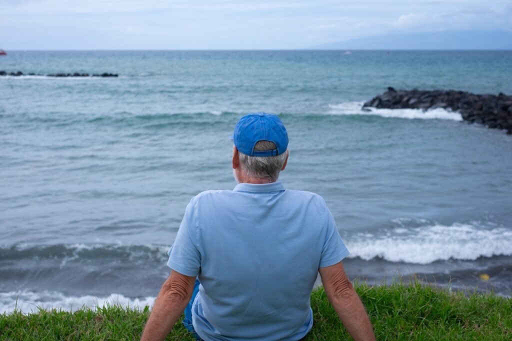 Rear view of senior man sitting on the grass at the beach in a cloudy day enjoying free time vacation or retirement. Relaxed man wearing blue cap looking at the horizon over sea