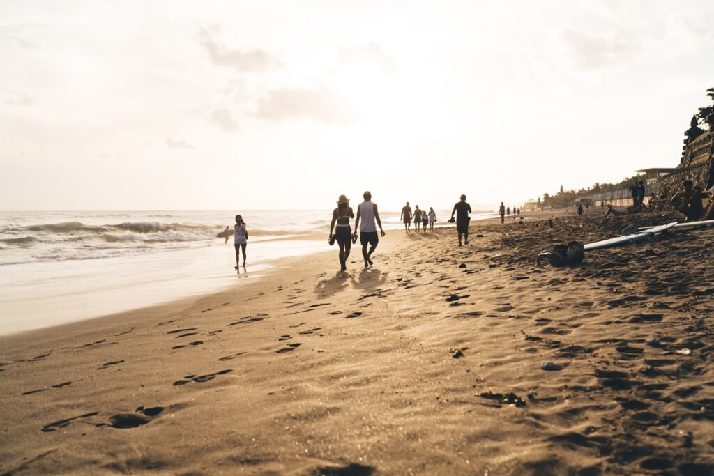 Back view of unrecognizable travel persons walking in relax on sun reflecting golden sandy beach near rolling in sea waves under bright sky