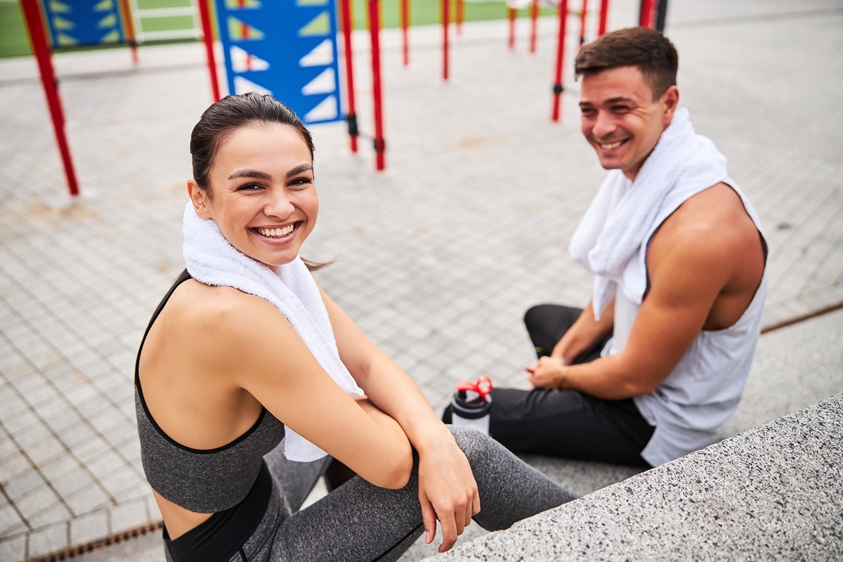 Top view waist up portrait of smiling woman with towel sitting near boyfriend after joint workout outdoors. Happy young couple relaxing after workout on sports ground