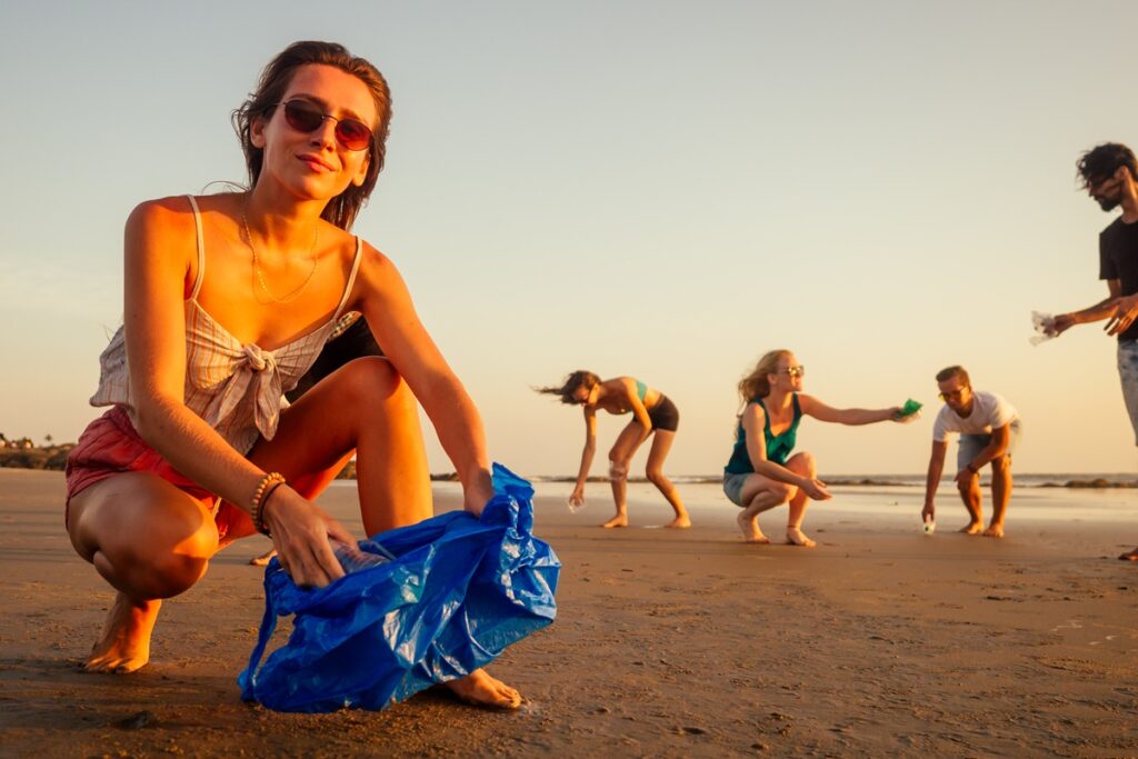 A woman kneels on the beach, a blue bag in her hands, as the sun sets over the horizon, casting a warm glow, ending the eco-tour in the French Riviera by giving back. 