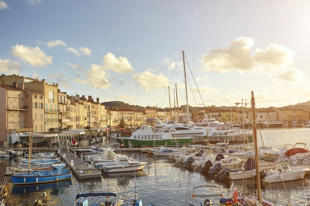 The natural, colourful harbour of Villefrance showcasing pastel coloured buildings, boats of all sizes, shapes and colours, and a pale blue sky with fluffy clouds crowning over the distant green mountains. View of harbour boats and waterfront, St Tropez, Cote d'Azur, France
