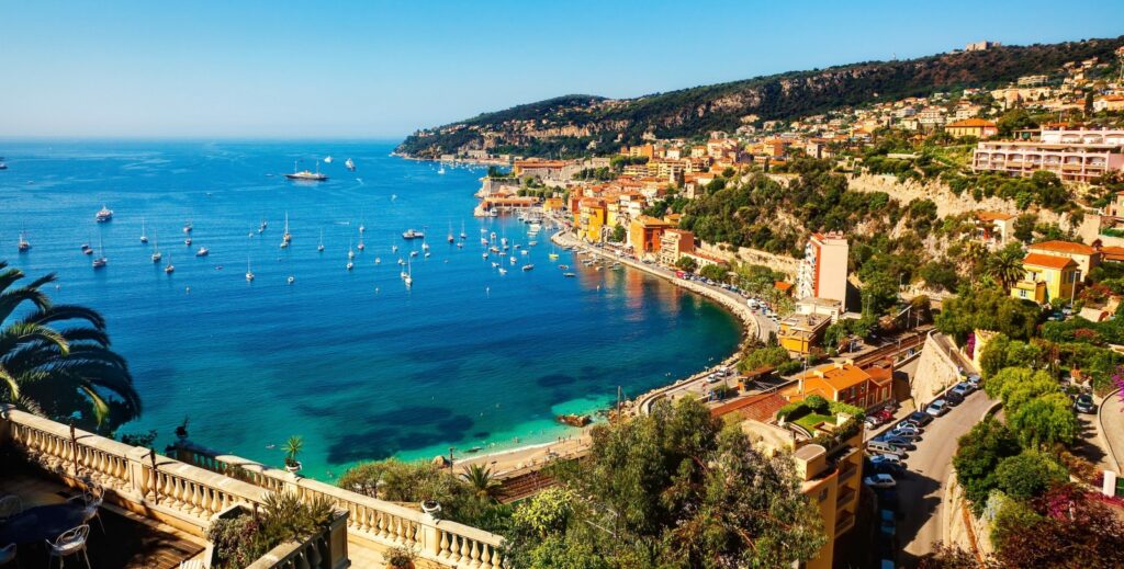 The vibrant coastal landscape of Villefranche-sur-Mer showcasing deep blue ocean, boats swaying, a vibrant sky, and pastel coloured buildings looking out onto the Mediterranean. Coastal landscape in France showcasing palm trees and boats against a vibrant sky.